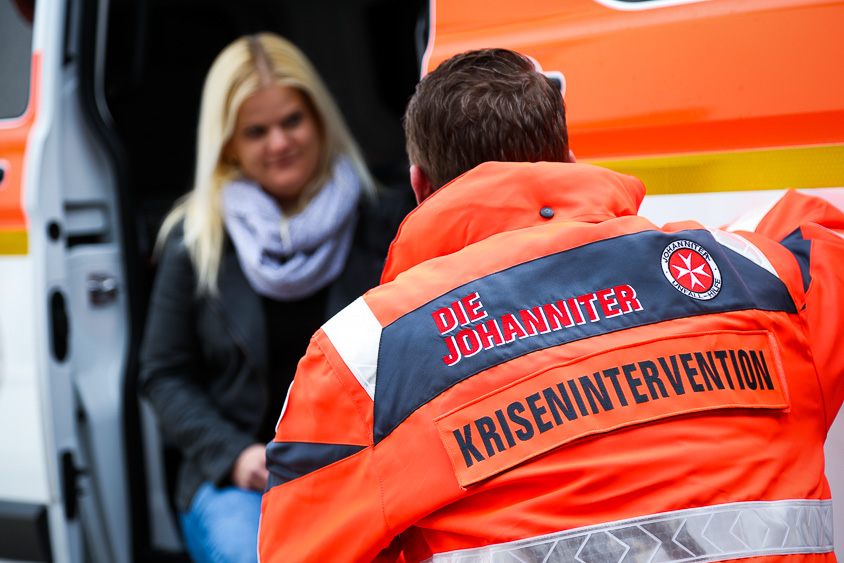 Ein ehrenamtlicher Helfer der Johanniter-Krisenintervention wendet sich einer Frau zu, um sie in einer emotionalen Ausnahmesituation zu unterstützen. Im Hintergrund ist ein Einsatzfahrzeug zu sehen.
