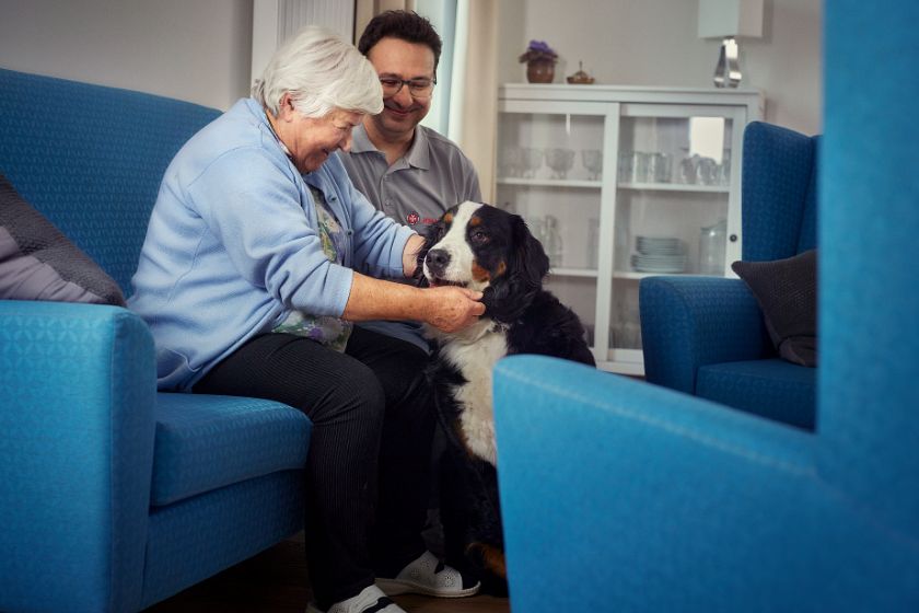 Wir sehen eine Pflegeperson und eine ältere Dame, die gemeinsam einen Hund streicheln.
