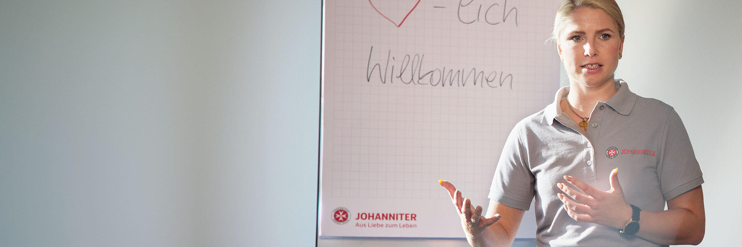 Johanniter-Mitarbeiterin im grauen Poloshirt spricht vor Flipchart mit Willkommensaufschrift.