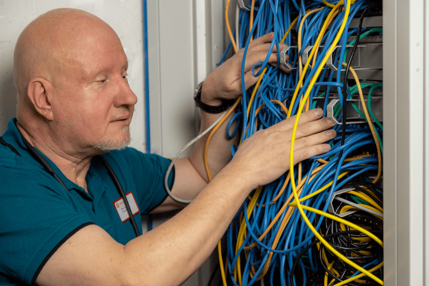 Ein kahlköpfiger Mann mit weißem Kinnbart vor einem Schrank mit vielen verschiedenfarbigen Kabeln