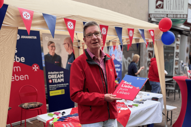 Ein Mann mit Brille steht vor einem Infostand der Johanniter. Er trägt eine rote Jacke und hält ein Schild mit der Aufschrift „Aus Liebe zum Leben“ in der Hand. Im Hintergrund sind ein Pavillon, Wimpelketten in Rot und Blau sowie weitere Informationsmaterialien der Johanniter zu sehen. 