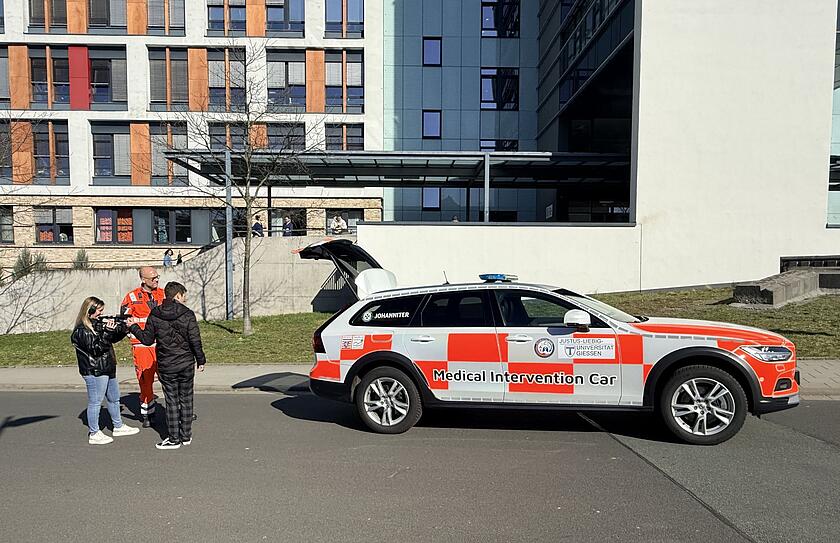 Drehszene vor UKGM-Gebäude mit GiMIC, zwei Personen Drehteam und Notfallsanitäter von der Johanniter-Unfall-Hilfe e.V.