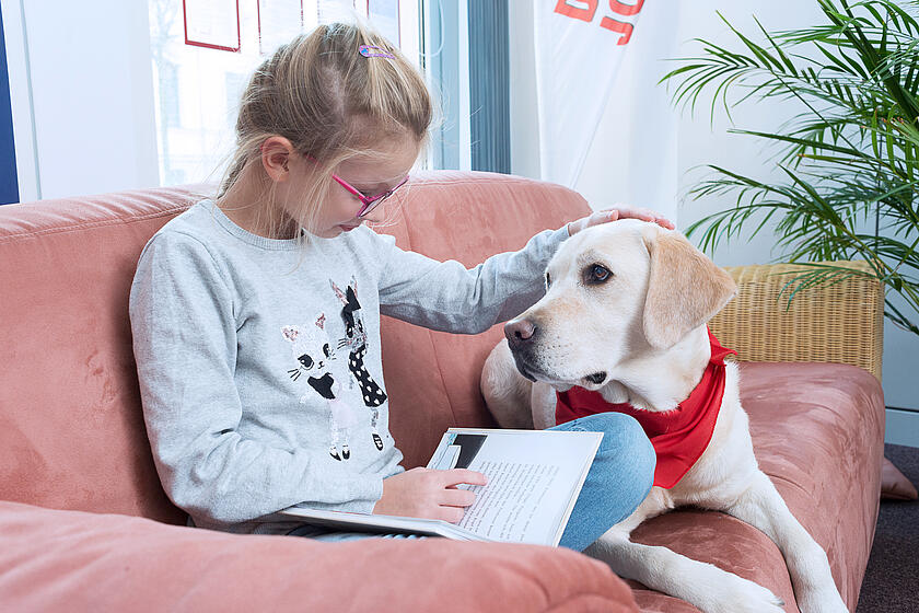 Ein Mädchen liest einem Lesehund etwas aus einem Buch vor und streichelt ihn dabei.