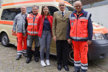 Fünf Ehrenamtliche der Johanniter stehen vor einem Einsatzfahrzeug. Alle tragen verschiedene Johanniter-Einsatz- oder Festkleidung und lächeln.