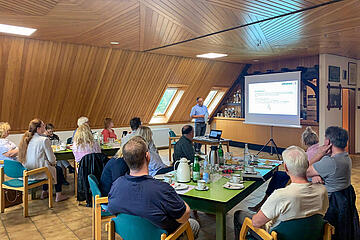 Im Gemeindehaus Tramm findet ein Grundlagenseminar statt. Zu sehen sind Ehrenamtliche, die an einem Tisch sitzen, und einer vortragenden Person vor einer Leinwand zuhören.