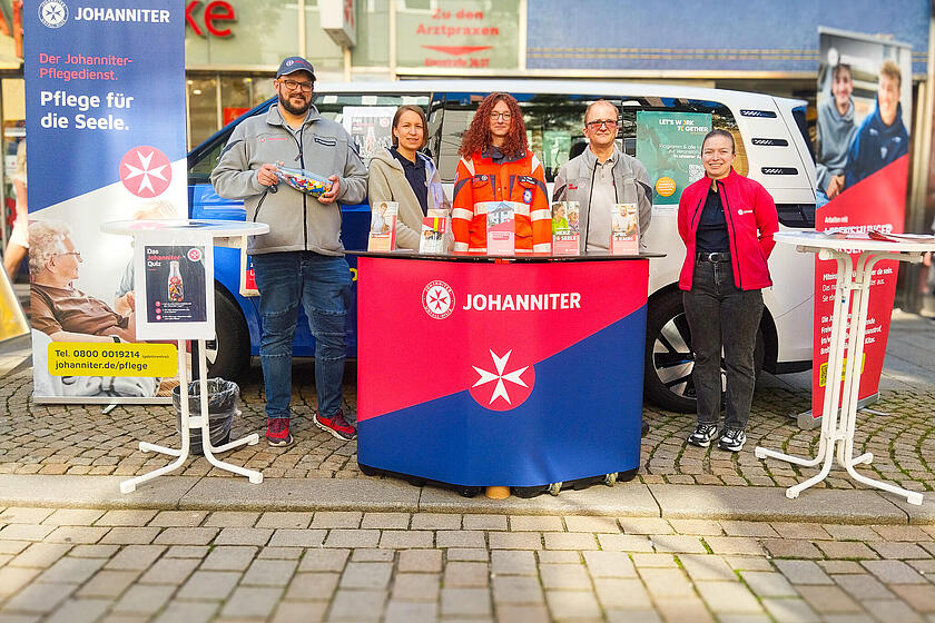 Frauen und Männer in Johanniter-Kleidung vor einem Messepult in einer Fußgängerzone. Rechts und links stehen Roll-Ups. Im Hintergrund steht ein Bulli.