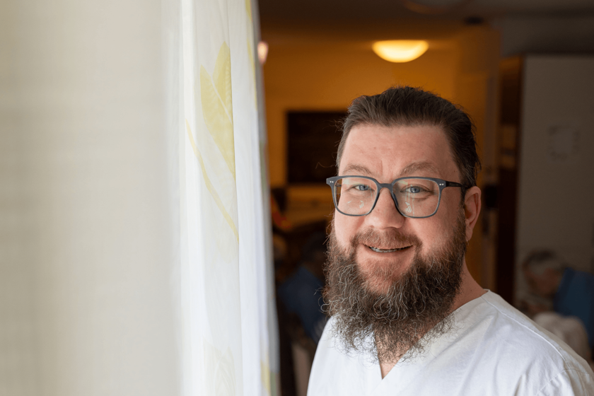 Ein Mann in weißer Arbeitskleidung steht am Fenster neben einem Vorhang, lächelt 