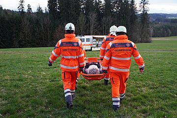 Vier Johanniter-Rettungskräfte in Einsatzkleidung tragen einen Patienten in einer Trage über eine Wiese.