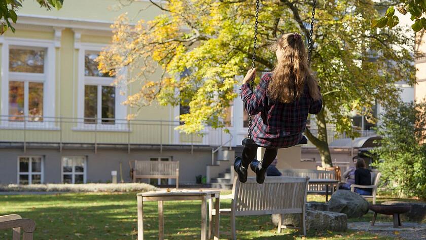 Person schaukelt im Garten der Klinik am Korso im Sonnenschein - zwei Personen unterhalten sich im Hintergrund