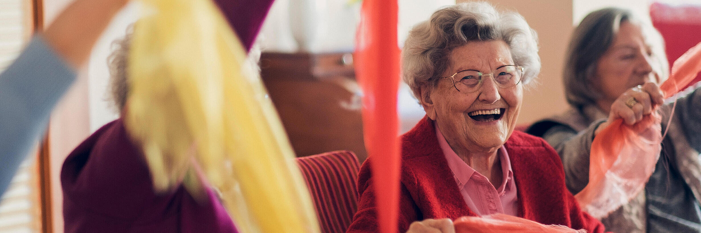 Lachende auf dem Stuhl sitzende Seniorinnen bewegen sich spielend mit einem bunten Tuch