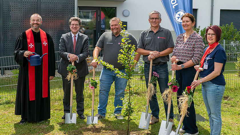 Ein Johanniter-Pfarrer und 5 weitere Johanniter stehen bei einer feierlichen Baumpflanzaktion vor einem Gebäude. Der Pfarrer hält eine Gießkanne in der Hand, die anderen halten jeweils einen geschmückten Spaten. Im Hintergrund ist eine Fahne, auf der "Johanniter" steht.