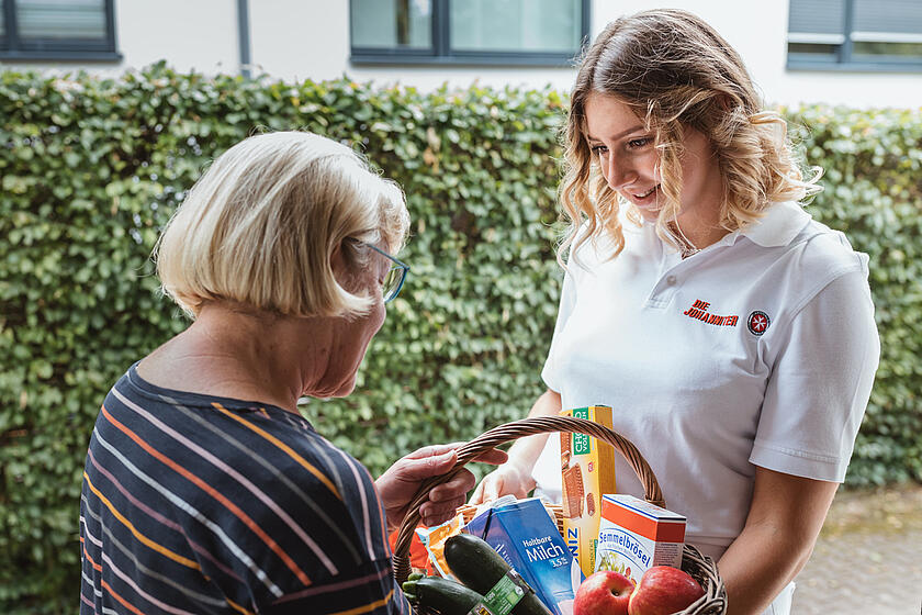 Mitarbeiterin vom mobilen sozialen Hilfdienst der Johanniter bringt einer Seniorin einen Korb mit Einkäufen an die Tür.