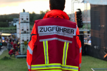 Ein ehrenamtlicher Helfer in Rückenansicht blickt auf ein Festivalgeschehen vor ihm an einem See. Er trägt eine Weste, die ihn als Zugführer ausweist.