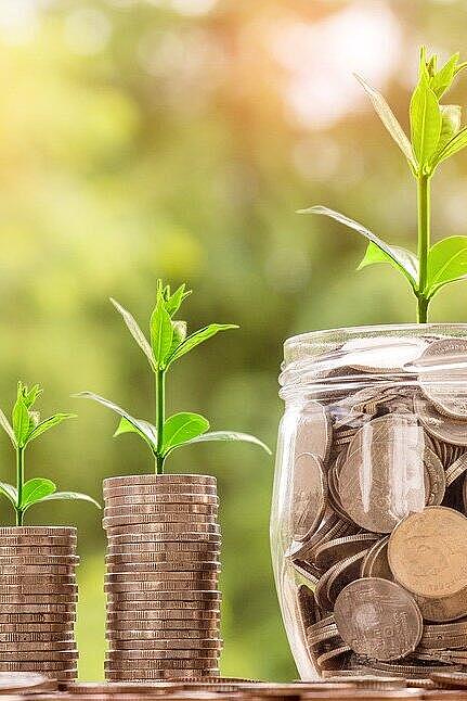 Gestapelte Münzen und ein Glas voller Geld, aus denen grüne Pflänzchen wachsen, symbolisieren finanzielles Wachstum.