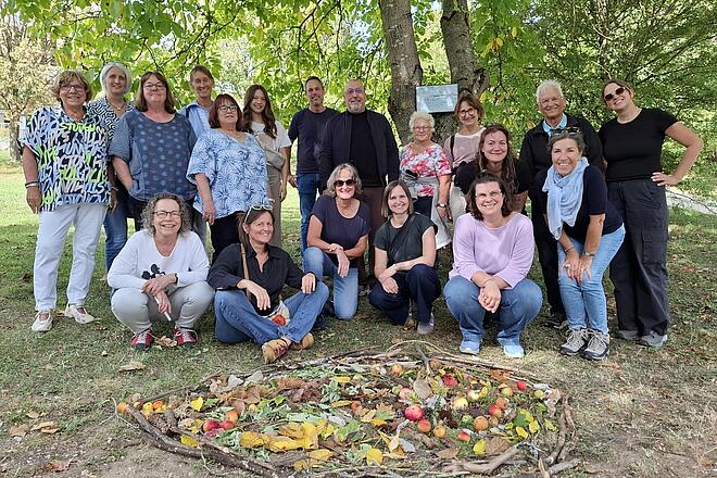 Gruppenfoto der Johanniter-Mitarbeitenden beim Lacrima-Mitarbeitendenwochenende unter Bäumen im Freien. Davor liegt ein herbstliches Mandala aus Ästen, Blättern, Tannenzapfen und Äpfeln.