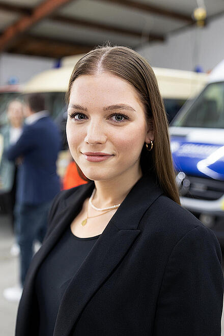 Junge Frau vor mehreren Einsatzfahrzeugen in einer Fahrzeughalle, Blick in die Kamera.
