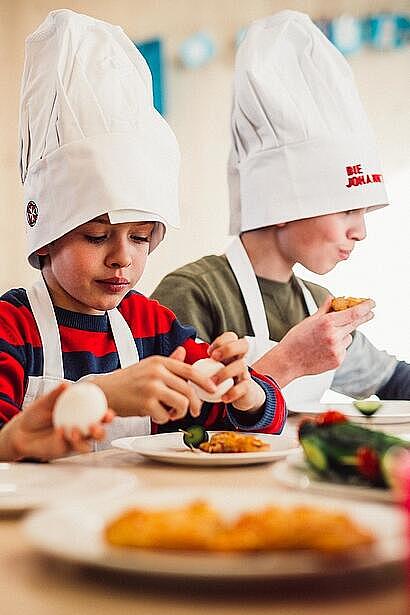 Drei Kinder in Kochmützen mit Johanniter-Logo sitzen am Tisch und bereiten Essen zu.