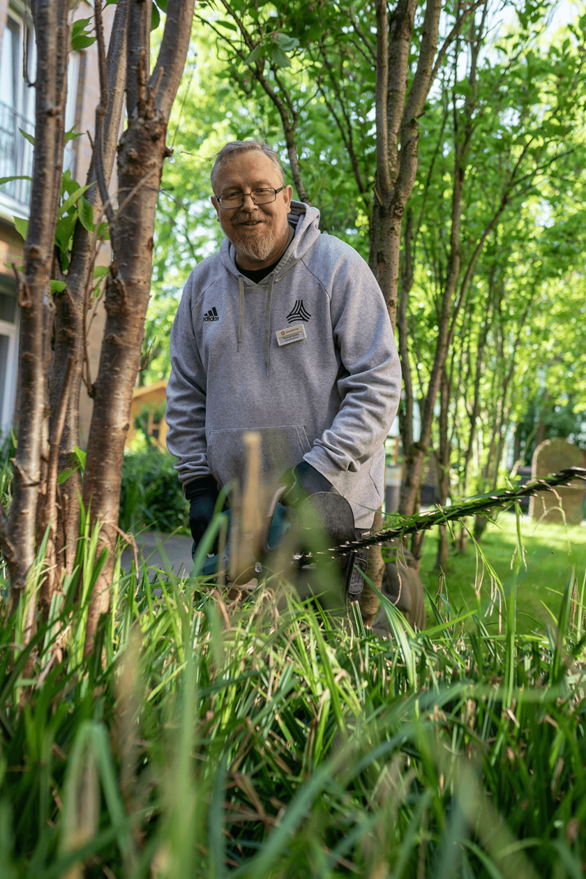 Ein Mann in grauer Arbeitskleidung schneidet im Garten mit einer Heckenschere Pflanzen