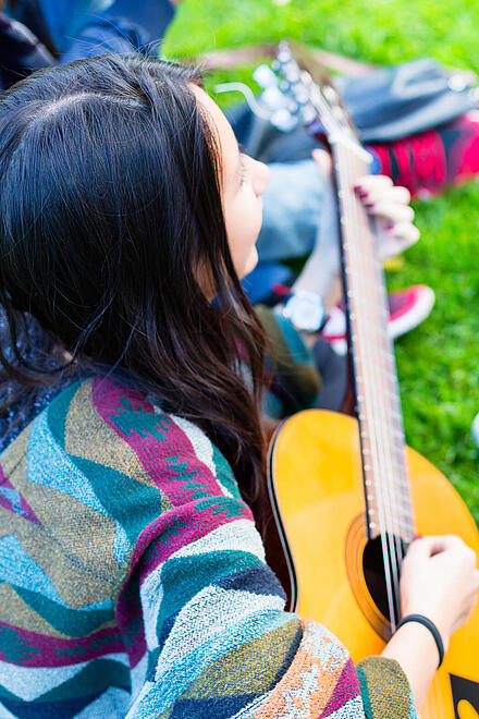 Eine Gruppe junger Menschen sitzt draußen auf grünem Gras um eine junge Frau mit Gitarre.