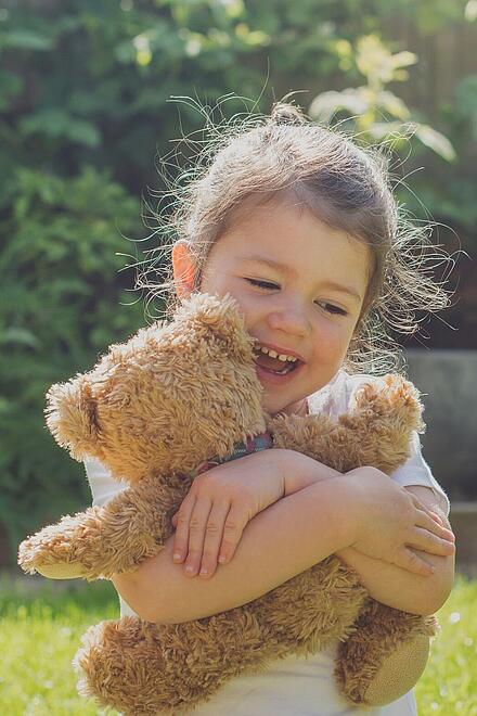 Ein lachendes Kind sitzt im Garten und umarmt fröhlich einen braunen Teddybären im Sonnenschein.