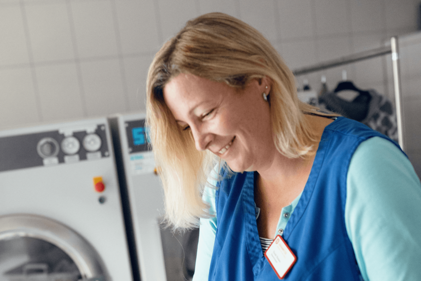 eine lächelnde blonde Frau in türkisem T-Shirt und blauem Kasack vor einer Reihe Waschmaschinen