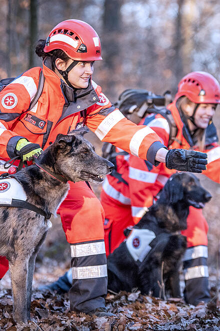 Zwei Johanniter in Einsatzkleidung mit ihren beiden Rettungshunden bei einem Sucheinsatz im Wald.