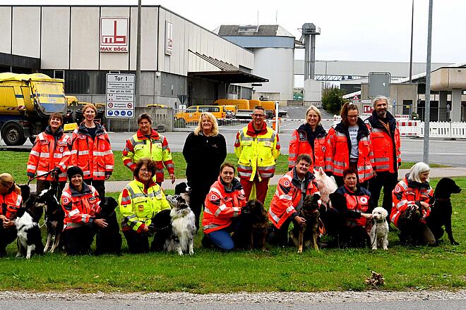 Gruppenfoto von Rettungshundeteams verschiedener Hilfsorganisationen mit ihren Hunden auf dem Gelände der Firma Max Bögl. Hundeführende in orangen und gelben Einsatzjacken knien gemeinsam mit ihren Hunden auf einer Wiese.