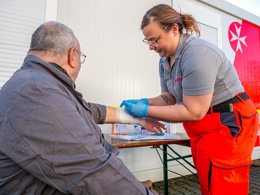 Eine Helferin der Johanniter beugt sich über den Arm eines älteren Mannes und legt einen Verband am Unterarm an.