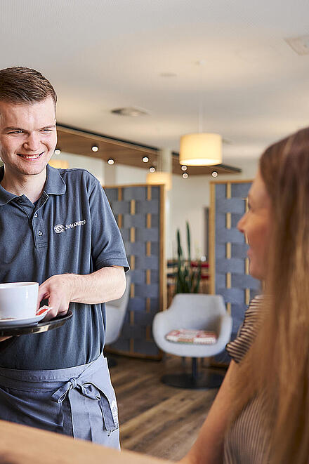 Includio Mitarbeitender serviert lächelnd einen Kaffee