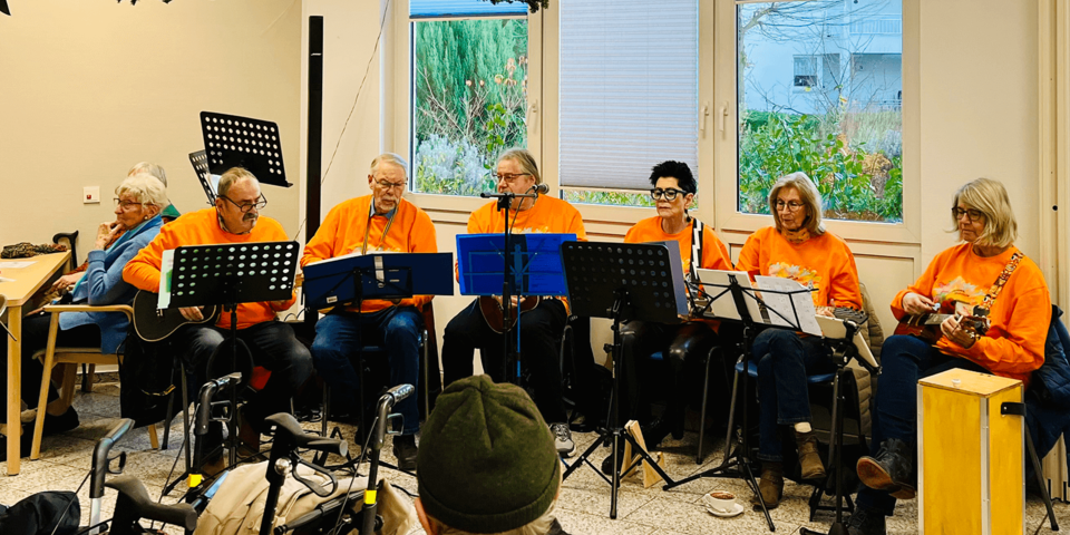 Eine Band mit mehreren Mitgliedern in orangefarbenen T-Shirts spielt gemeinsam auf Ukulelen und anderen Instrumenten in einem festlich geschmückten Raum; vor ihnen sitzen Zuschauende, einige mit Rollator.