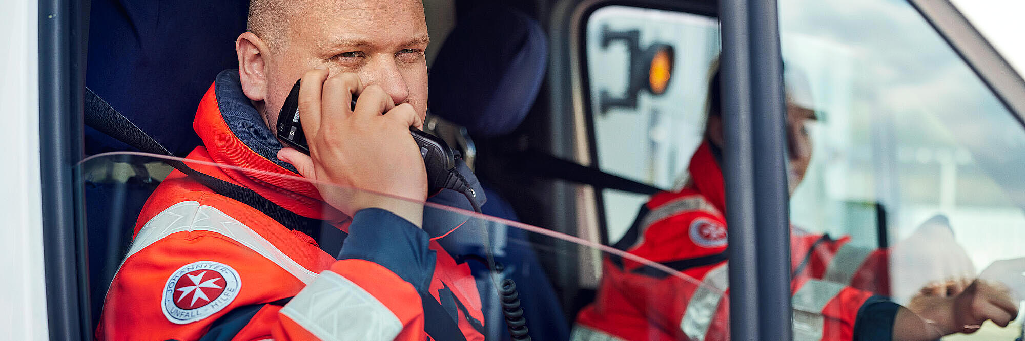 Zwei Johanniter sitzen in Einsatzkleidung in einem Einsatzfahrzeug und einer von ihnen spricht in ein Funkgerät.